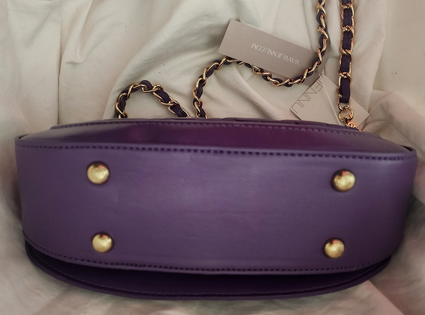 A view of the bottom of the purple vegan leather Taipei shoulder bag with gold-tone hardware, featuring four gold stud feet and a removable, gold and purple chain strap, resting on a white fabric background.