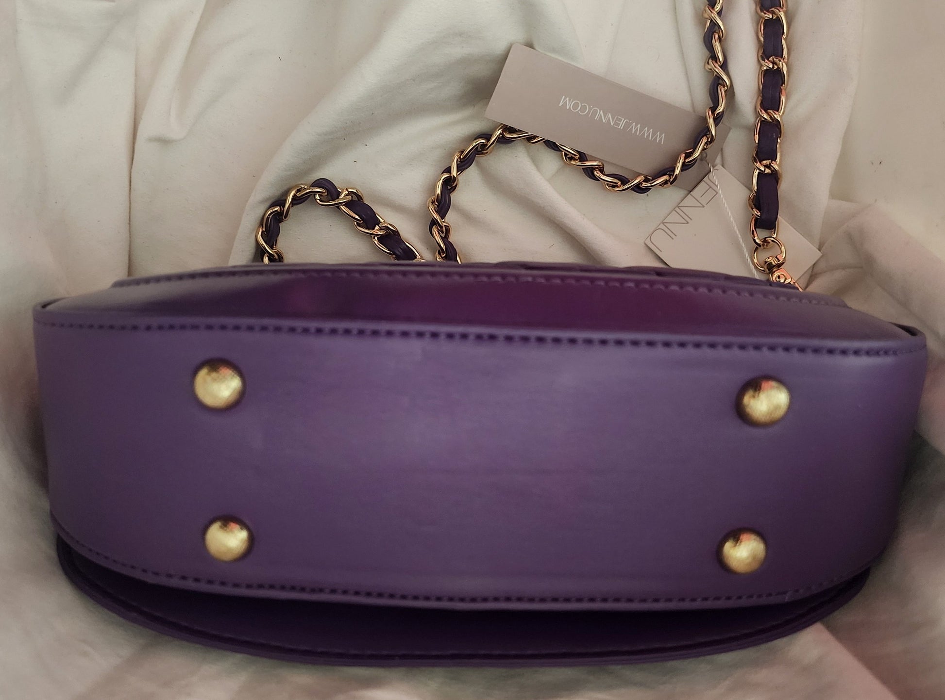 A view of the bottom of the purple vegan leather Taipei shoulder bag with gold-tone hardware, featuring four gold stud feet and a removable, gold and purple chain strap, resting on a white fabric background.