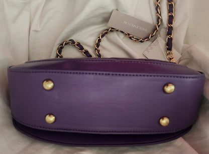 A view of the bottom of the purple vegan leather Taipei shoulder bag with gold-tone hardware, featuring four gold stud feet and a removable, gold and purple chain strap, resting on a white fabric background.