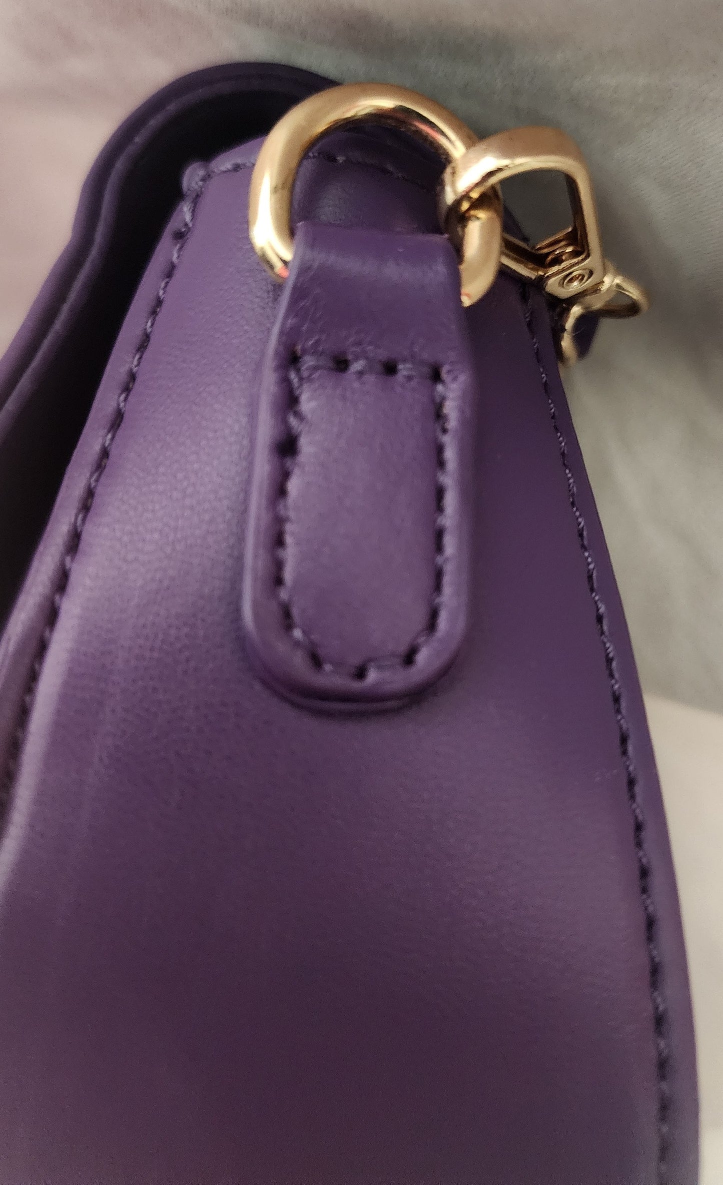 Close-up of the purple Taipei vegan leather shoulder purse showing a stitched tab, a gold-tone metal ring, and a gold lobster claw clasp for the removable shoulder strap on a soft light gray background.