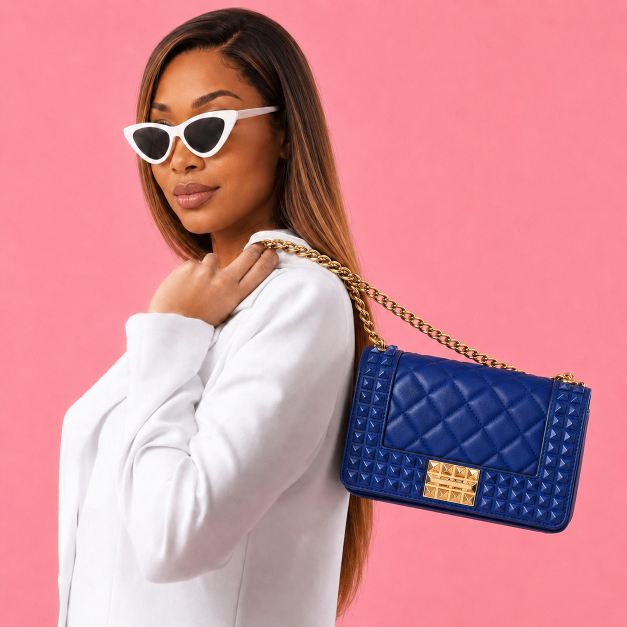 Purse-illa showcases the Donna Studded Jelly Chain Crossbody Bag worn over the shoulder. The cobalt blue quilted bag features pyramid studs, a gold chain strap, and bold hardware, creating a sophisticated statement accessory against a warm golden backdrop.