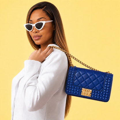Purse-illa showcases the Donna Studded Jelly Chain Crossbody Bag worn over the shoulder. The cobalt blue quilted bag features pyramid studs, a gold chain strap, and bold hardware, creating a sophisticated statement accessory against a warm golden backdrop.