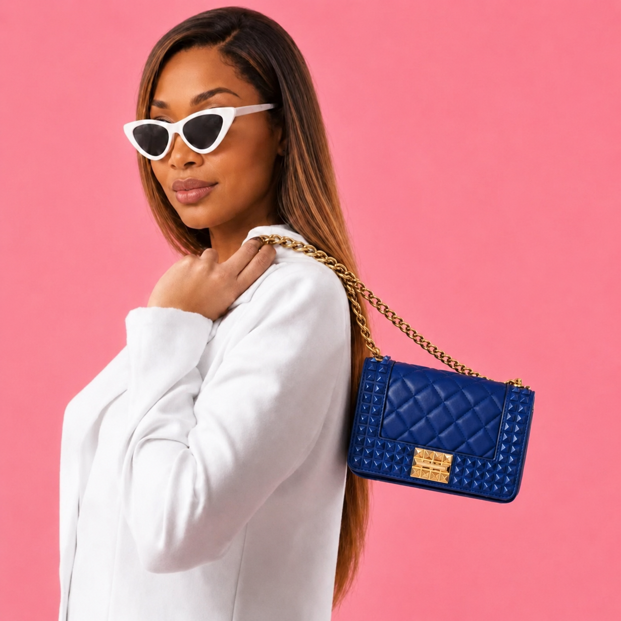 Purse-illa showcases the Donna Studded Jelly Chain Crossbody Bag worn over the shoulder. The cobalt blue quilted bag features pyramid studs, a gold chain strap, and bold hardware, creating a sophisticated statement accessory against a warm golden backdrop.