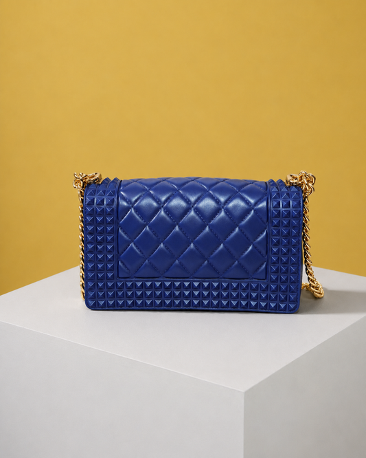 The back view of the Donna Studded Jelly Chain Crossbody Bag showcases a cobalt blue quilted design with pyramid studs, structured edges, and a gold chain strap. The bag rests on a white pedestal against a warm golden background for a luxury editorial presentation.