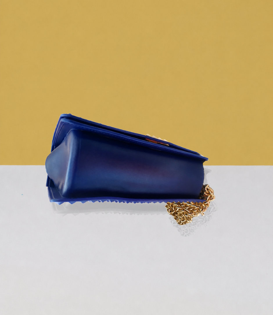 The bottom view of the Donna Studded Jelly Chain Crossbody Bag highlights its smooth cobalt blue jelly vegan leather construction, structured base, studded edges, and gold chain detail. The compact silhouette is displayed on a white pedestal against a warm golden backdrop.