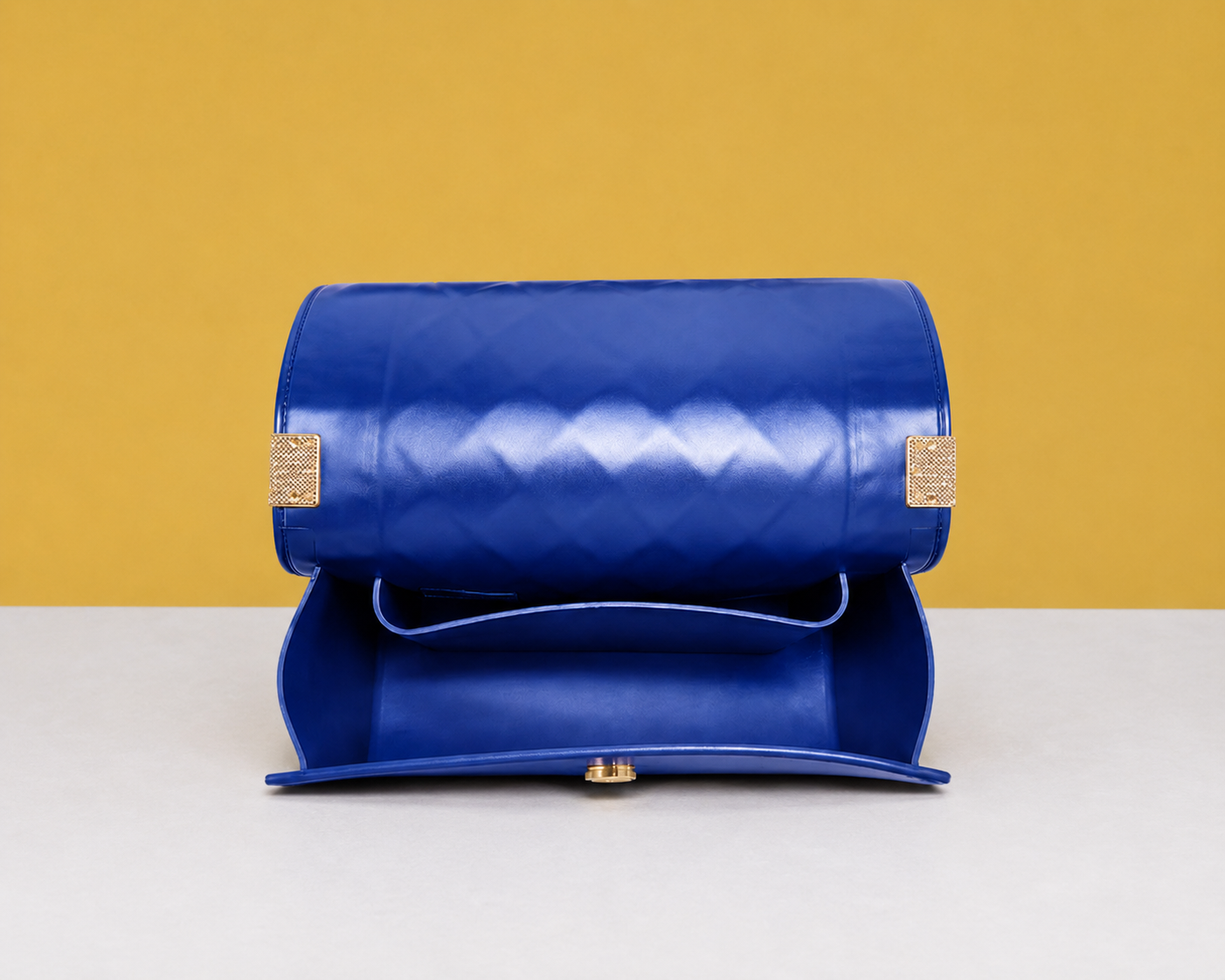 The opened Donna Studded Jelly Chain Crossbody Bag reveals its spacious interior and smooth flap design. The cobalt blue quilted exterior, gold accents, and structured silhouette are showcased on a white pedestal against a warm golden editorial backdrop.