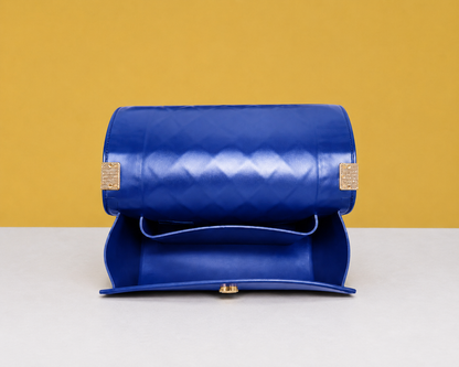 The opened Donna Studded Jelly Chain Crossbody Bag reveals its spacious interior and smooth flap design. The cobalt blue quilted exterior, gold accents, and structured silhouette are showcased on a white pedestal against a warm golden editorial backdrop.