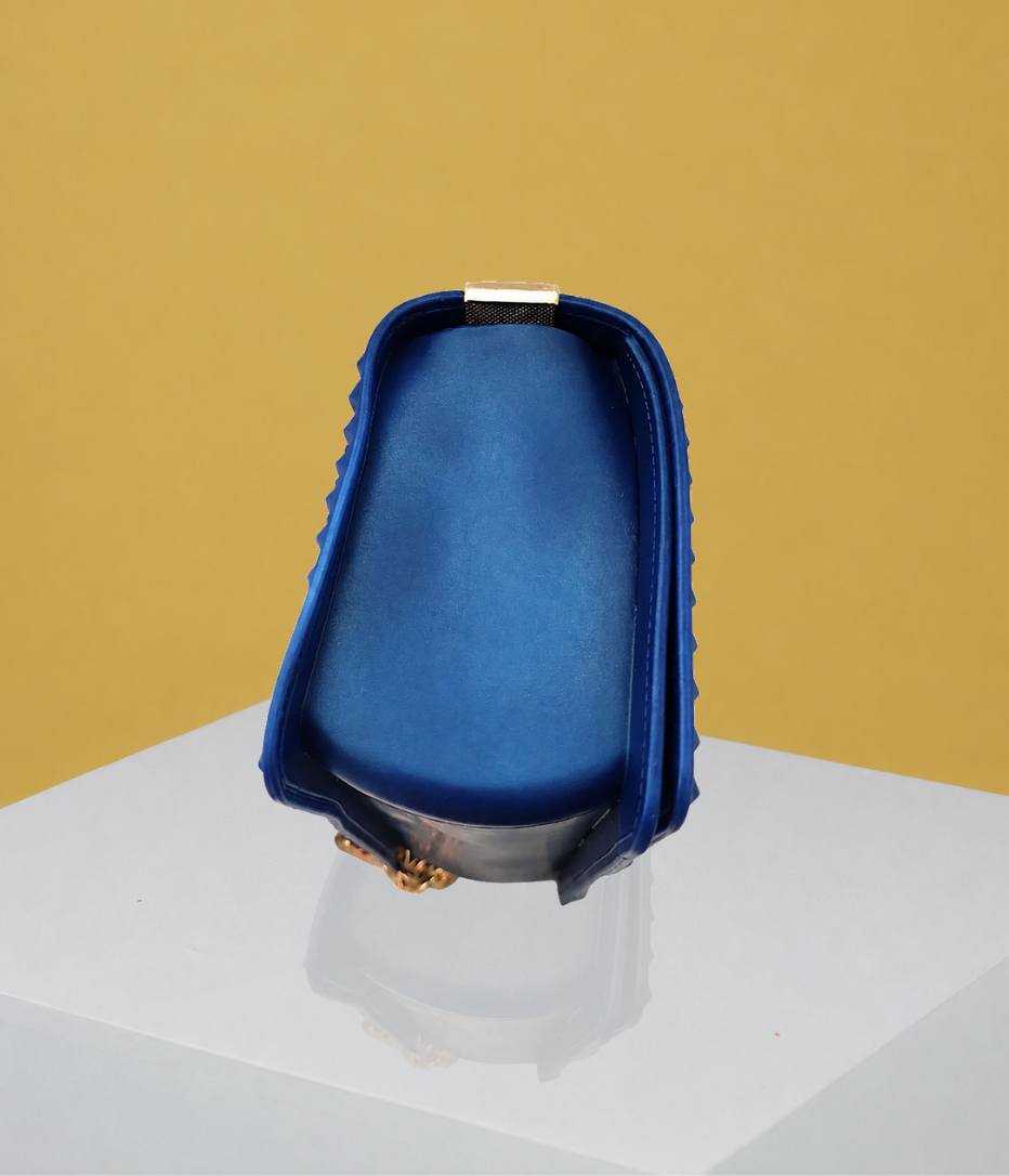 The side view of the Donna Studded Jelly Chain Crossbody Bag highlights its structured silhouette, cobalt blue finish, raised studded edges, and compact profile. Displayed on a white pedestal, the bag is presented against a warm golden editorial backdrop.