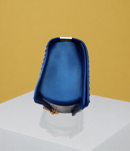 The side view of the Donna Studded Jelly Chain Crossbody Bag highlights its structured silhouette, cobalt blue finish, raised studded edges, and compact profile. Displayed on a white pedestal, the bag is presented against a warm golden editorial backdrop.