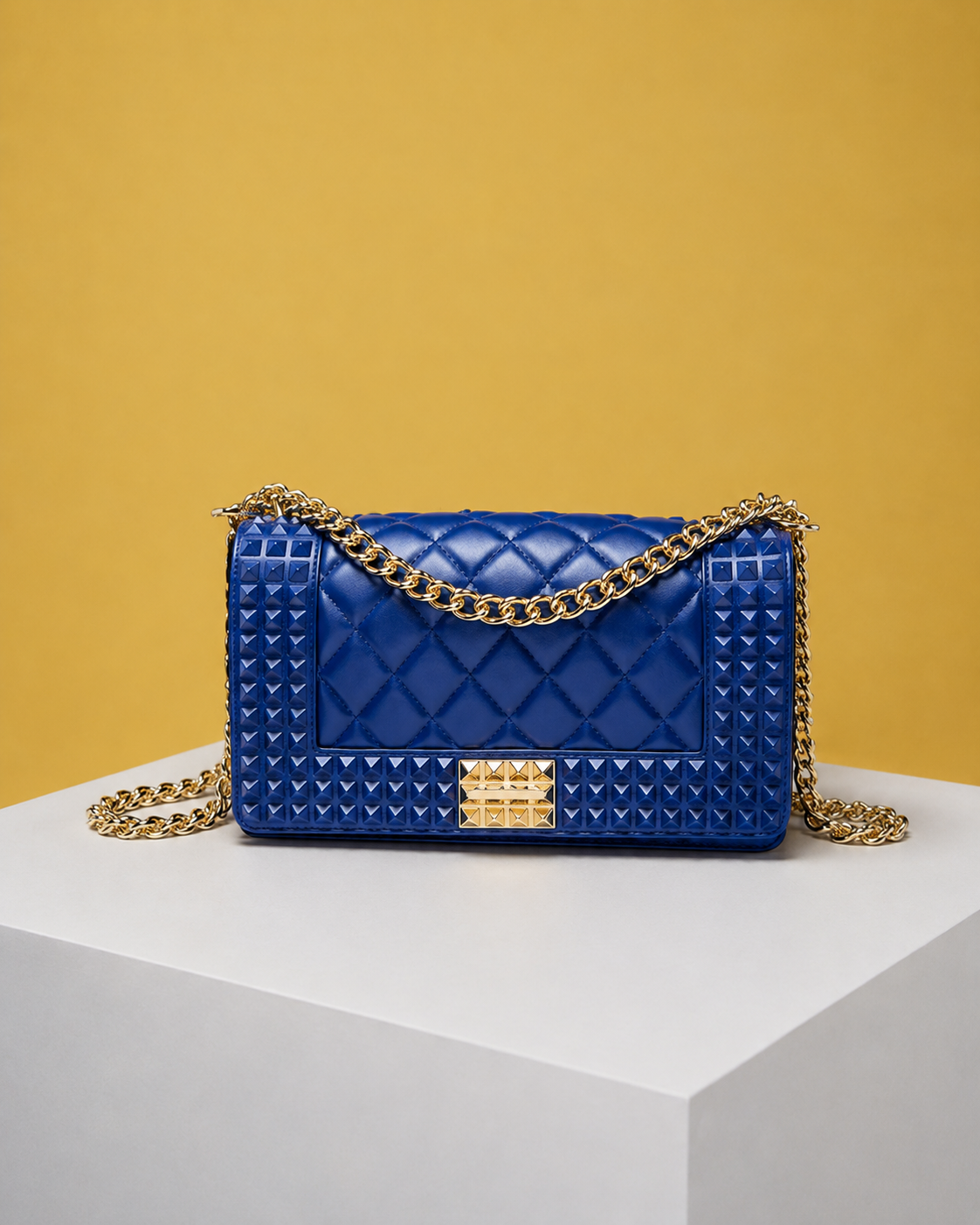 The front view of the Donna Studded Jelly Chain Crossbody Bag showcases cobalt blue jelly vegan leather, quilted detailing, pyramid studs, and a gold chain strap. Displayed on a white pedestal, the statement bag creates a luxurious editorial presentation.